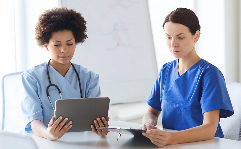 Two female medical professionals, seated at a round table, study an electronic pad with information.
