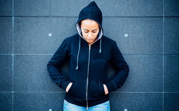 Young black girl in hoodie and jeans leans against wall, eyes down