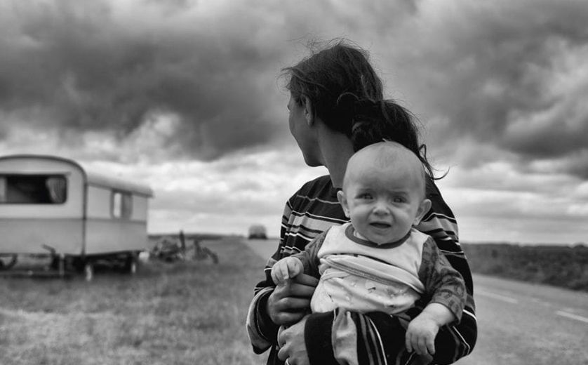 A woman holds an infant facing camera and looks back over her shoulder next to a deserted road