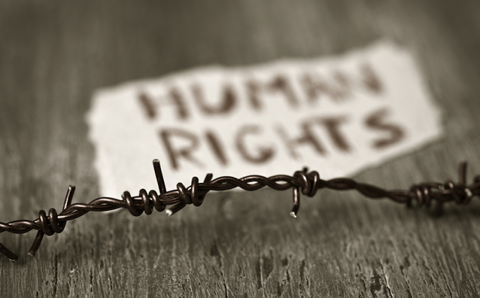 Black and white photo; barbed wire on table in foreground; paper with words 'human rights' in background.