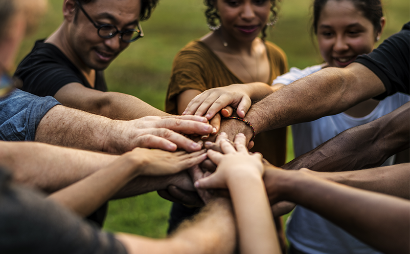 A group of people in a park, from differing ethnicities stand in a circle, hands in.