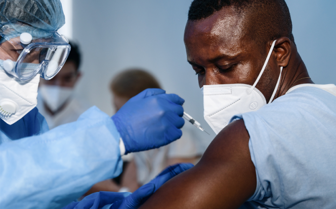A medical professional delivers a vaccine shot to an African (American) man; both are masked.
